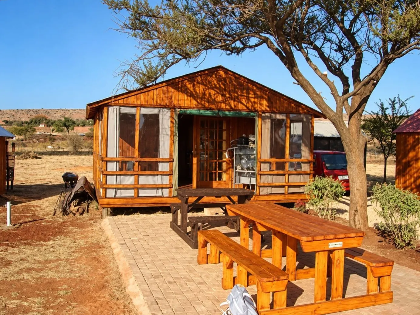 A timber bar hut with screened sides, a railed veranda and a glass-panelled door, with picnic tables on a paved area under a thorn tree
